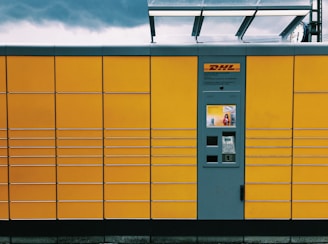 A yellow package locker system with a grey central panel featuring a screen and several compartments. The brand logo is visible above the panel. The structure has horizontal lines creating rectangular sections. The top of the locker has a transparent overhang, and the sky is overcast with dark clouds.