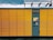 A yellow package locker system with a grey central panel featuring a screen and several compartments. The brand logo is visible above the panel. The structure has horizontal lines creating rectangular sections. The top of the locker has a transparent overhang, and the sky is overcast with dark clouds.