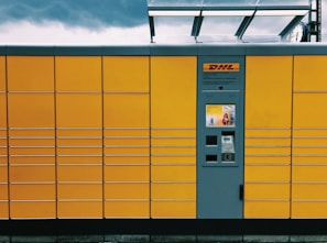 A yellow package locker system with a grey central panel featuring a screen and several compartments. The brand logo is visible above the panel. The structure has horizontal lines creating rectangular sections. The top of the locker has a transparent overhang, and the sky is overcast with dark clouds.