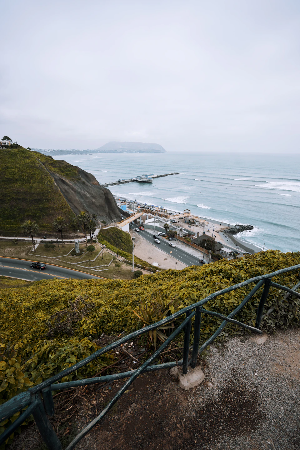 The image depicts a coastal landscape with a cliff overlooking the ocean, featuring a pier extending into the water and several buildings scattered across the shoreline. The sky is overcast, casting a soft light on the scene below. In the foreground, there are lush green bushes that add a touch of color to the otherwise muted palette of the image.

In the background, cars can be seen driving along the road leading up to the cliff, and a few people are visible walking near the pier or enjoying the view from the top of the hill. The overall scene conveys a sense of tranquility and natural beauty, with the ocean stretching out into the horizon under an overcast sky.