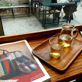 A cozy caf&eacute; setting featuring a wooden table with a tray holding a glass and a small pitcher of tea. An open magazine displays luxury items such as a handbag and leather shoes. Beyond the window, an outdoor seating area with bench and table can be seen. Two people are seated at a nearby table.