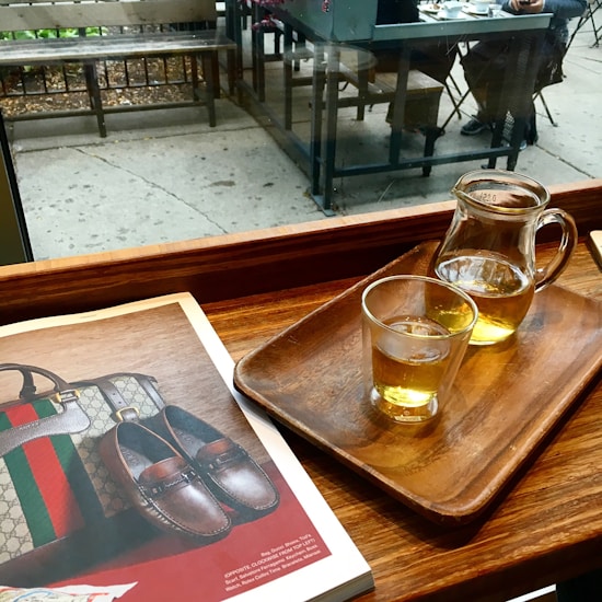 A cozy caf&eacute; setting featuring a wooden table with a tray holding a glass and a small pitcher of tea. An open magazine displays luxury items such as a handbag and leather shoes. Beyond the window, an outdoor seating area with bench and table can be seen. Two people are seated at a nearby table.
