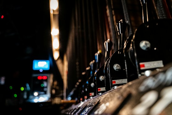 Row of industrial control levers with metal handles and labels aligned on a panel, with a background displaying soft focus lights and electronic screens, suggesting a mechanical or backstage setting.