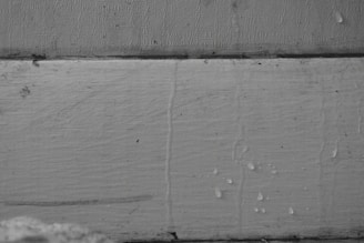 A close-up view of a textured, painted, wooden surface with visible cracks and small water droplets. The wood appears aged, with some areas darker due to moisture. The background is an unevenly painted section separated by a horizontal line at the top.