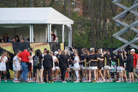 A group of athletes in sports uniforms is gathered on what appears to be a lacrosse field. They are standing in two groups, some wearing white uniforms and some wearing dark uniforms. There is a tent with people inside, possibly officials or commentators, overlooking the field in the background. Extensive greenery and trees are visible behind the tent.