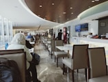 A modern airport lounge with sleek, comfortable seating and a long row of tables. The ceiling is curved with recessed lighting, and a flat-screen monitor displays information on the wall. Several people are seated and working or resting, while a few staff members in uniform attend to the area.
