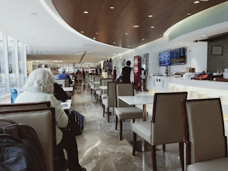 A modern airport lounge with sleek, comfortable seating and a long row of tables. The ceiling is curved with recessed lighting, and a flat-screen monitor displays information on the wall. Several people are seated and working or resting, while a few staff members in uniform attend to the area.