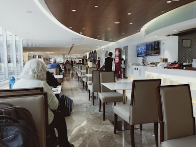 A modern airport lounge with sleek, comfortable seating and a long row of tables. The ceiling is curved with recessed lighting, and a flat-screen monitor displays information on the wall. Several people are seated and working or resting, while a few staff members in uniform attend to the area.