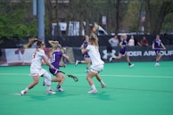 A group of female athletes is engaged in a competitive lacrosse game on a green field. Two players in white jerseys are actively challenging a player in a purple jersey who is holding a lacrosse stick. Other players and people can be seen in the background near the sidelines.