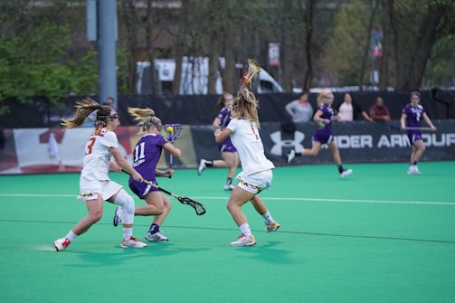 A group of female athletes is engaged in a competitive lacrosse game on a green field. Two players in white jerseys are actively challenging a player in a purple jersey who is holding a lacrosse stick. Other players and people can be seen in the background near the sidelines.