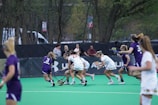 A group of female athletes play lacrosse on a green artificial turf field. The players are wearing uniforms, with some in white and others in purple. Spectators are visible in the background behind a black fence, and trees line the edge of the field.