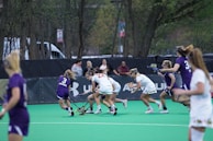 A group of female athletes play lacrosse on a green artificial turf field. The players are wearing uniforms, with some in white and others in purple. Spectators are visible in the background behind a black fence, and trees line the edge of the field.