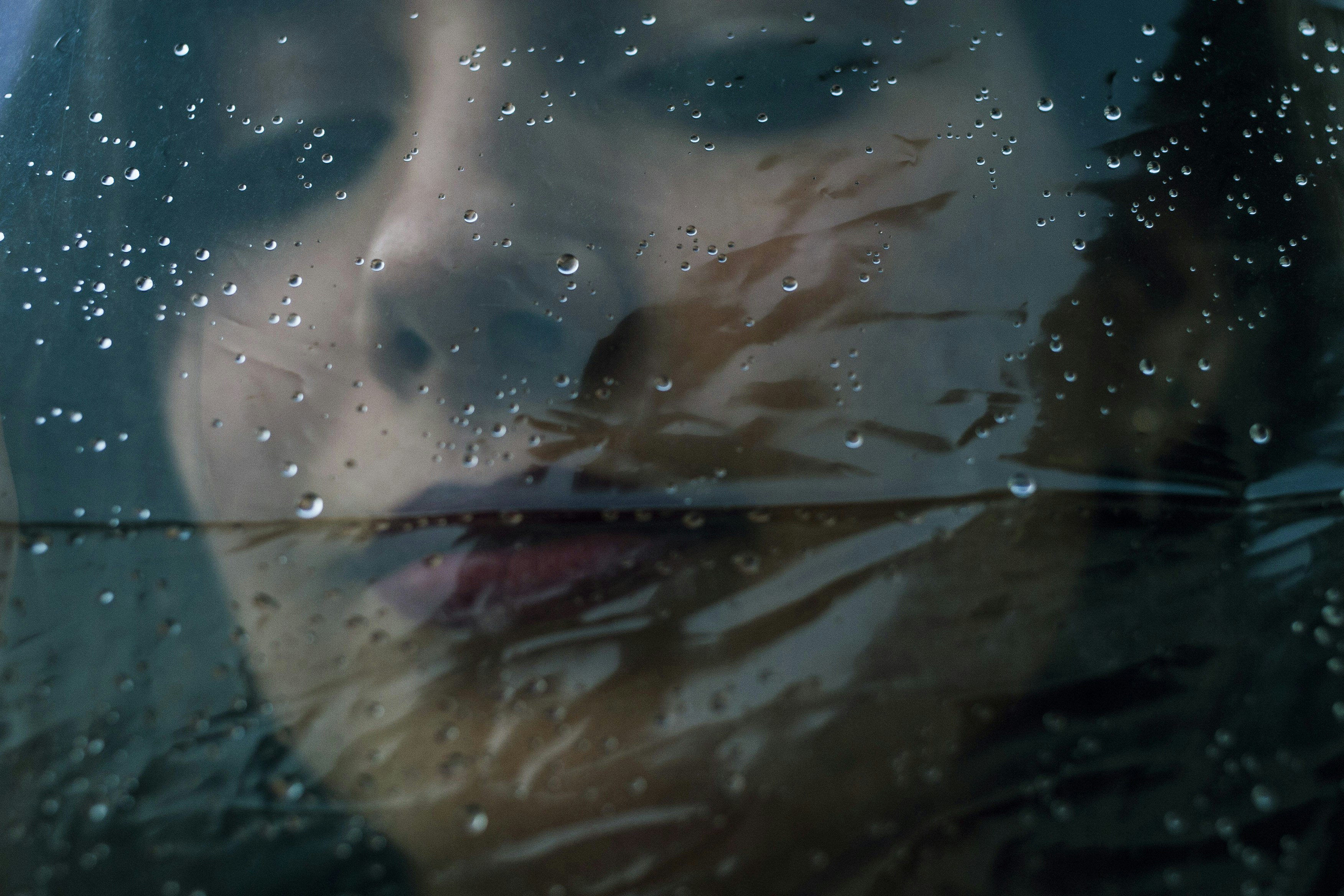 Dark, close up photo of a woman's face, looking down, behind a sheet of clear plastic covered with water droplets like rain 