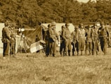 A group of soldiers stands in formation on a grassy field, wearing vintage military uniforms and helmets. Behind them are several canvas tents, indicating a temporary campsite. The background is filled with dense trees, and the image has a sepia tone, giving a historical or nostalgic feel.
