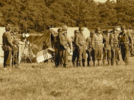 A group of soldiers stands in formation on a grassy field, wearing vintage military uniforms and helmets. Behind them are several canvas tents, indicating a temporary campsite. The background is filled with dense trees, and the image has a sepia tone, giving a historical or nostalgic feel.