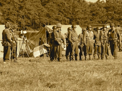 A group of soldiers stands in formation on a grassy field, wearing vintage military uniforms and helmets. Behind them are several canvas tents, indicating a temporary campsite. The background is filled with dense trees, and the image has a sepia tone, giving a historical or nostalgic feel.