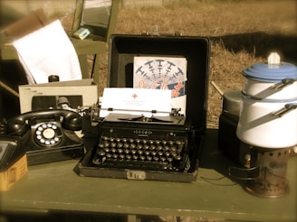 A vintage setup features an old-fashioned typewriter with a sheet of paper inserted, accompanied by a rotary dial telephone. There are various items on a table, including an enamel pot with a lid and a metal stand. The background has a nostalgic air, with an aged photo and a poster depicting airplanes.