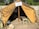 A vintage military tent with an open entrance is set up in an outdoor area. The tent is made of brown fabric and covered partially by camouflage netting. Inside the tent, there are various items including clothing, bottles, and a bedroll, indicating a temporary living setup. A sign above the entrance suggests a World War II German military context. Surrounding the tent are military gear and equipment such as jerry cans and bags.