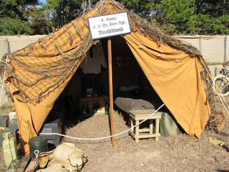 A vintage military tent with an open entrance is set up in an outdoor area. The tent is made of brown fabric and covered partially by camouflage netting. Inside the tent, there are various items including clothing, bottles, and a bedroll, indicating a temporary living setup. A sign above the entrance suggests a World War II German military context. Surrounding the tent are military gear and equipment such as jerry cans and bags.