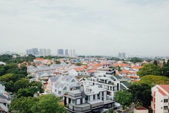 A sprawling suburban area with numerous houses featuring orange and gray roofs. The houses are surrounded by abundant greenery with tall trees interspersed throughout. In the background, there is a cluster of tall, modern buildings that indicate an urban skyline.