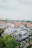 Aerial view of a suburban neighborhood featuring modern and traditional houses with red and gray roofs. Tall buildings in the distant background indicate a nearby cityscape. Lush green trees are scattered throughout, adding greenery to the area.