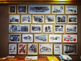 A collection of framed vintage motorcycle photographs and advertisements is displayed on a wall. The images feature different models of motorcycles, some with riders and others highlighting the design and engineering aspects. The photographs also include scenes of people interacting with the motorcycles and various promotional posters. Below the frames, there is a wooden surface with stacks of paper and a yellow folder.