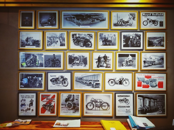 A collection of framed vintage motorcycle photographs and advertisements is displayed on a wall. The images feature different models of motorcycles, some with riders and others highlighting the design and engineering aspects. The photographs also include scenes of people interacting with the motorcycles and various promotional posters. Below the frames, there is a wooden surface with stacks of paper and a yellow folder.