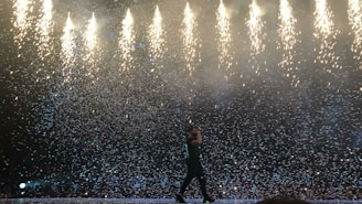 A performer on stage surrounded by a dramatic display of falling confetti and fireworks. The bright lights above contrast with the dark silhouette of the individual, creating a dynamic and energetic atmosphere.