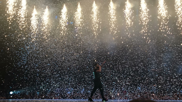 A performer on stage surrounded by a dramatic display of falling confetti and fireworks. The bright lights above contrast with the dark silhouette of the individual, creating a dynamic and energetic atmosphere.