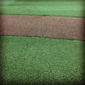 A textured surface featuring a path-like strip of coarse brown gravel running horizontally through two areas of vibrant green artificial grass.