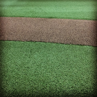 A textured surface featuring a path-like strip of coarse brown gravel running horizontally through two areas of vibrant green artificial grass.