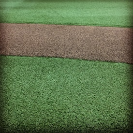 A textured surface featuring a path-like strip of coarse brown gravel running horizontally through two areas of vibrant green artificial grass.