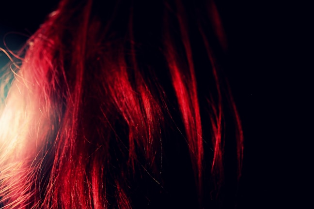 A close-up of vibrant red hair strands illuminated against a dark background, creating a striking contrast with the dark surroundings.
