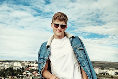 A person wearing sunglasses and a denim jacket stands confidently against a backdrop of a cityscape and a cloudy sky.