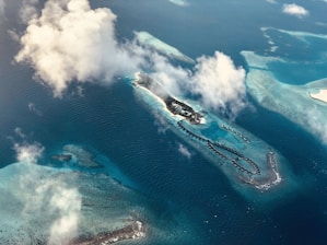 An aerial view of a tropical island surrounded by turquoise waters and coral reefs. There are overwater bungalows extending out into the sea from the island. Fluffy white clouds partially obscure the scene.
