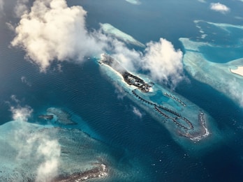 An aerial view of a tropical island surrounded by turquoise waters and coral reefs. There are overwater bungalows extending out into the sea from the island. Fluffy white clouds partially obscure the scene.