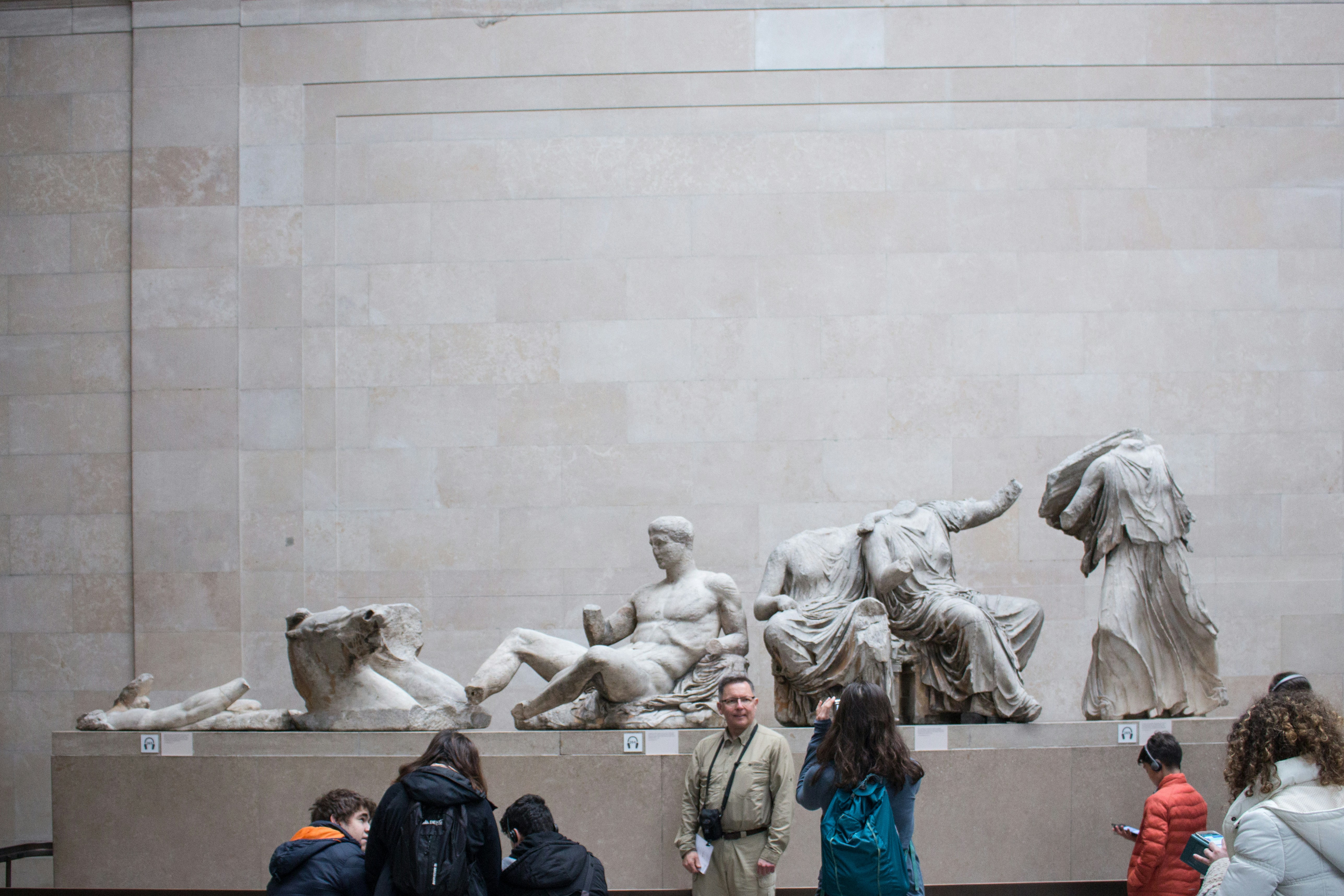 Creek Parthenon marble sculptures mounted along a limestone wall, with museum visitors walking past and reading exhibit text.