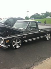 A classic black pickup truck with chrome wheels is parked on a paved surface. The truck is well-polished and appears to be in excellent condition, likely a vintage or custom model. Partially open hood suggests it might be on display at a car event. In the background, other vehicles and people are visible in a parking lot setting, with grassy areas and trees in the distance under an overcast sky.