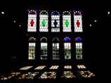 A series of large stained glass windows with elaborate geometric patterns. The glass panels feature vivid colors including red, blue, and green, creating patterns that project onto the floor. The windows are framed by a dark interior environment, enhancing the contrast between the colorful glass and the surrounding shadows.