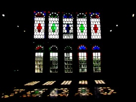 A series of large stained glass windows with elaborate geometric patterns. The glass panels feature vivid colors including red, blue, and green, creating patterns that project onto the floor. The windows are framed by a dark interior environment, enhancing the contrast between the colorful glass and the surrounding shadows.