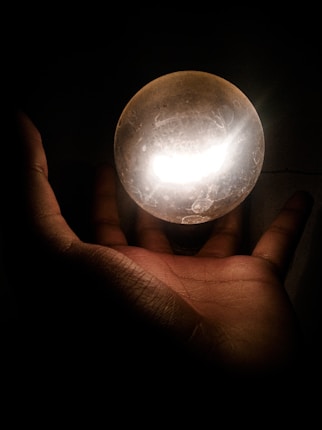A hand is shown holding a glowing, crystal-like sphere in a dark environment. The light from the sphere illuminates the hand and the immediate surroundings with an ethereal glow.