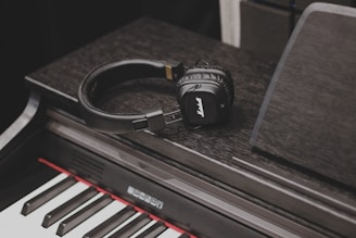 A pair of black headphones rests on an electronic keyboard. The keys of the keyboard are clearly visible, showing a combination of black and white keys with a red accent above the key bed.