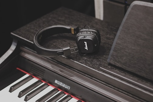 A pair of black headphones rests on an electronic keyboard. The keys of the keyboard are clearly visible, showing a combination of black and white keys with a red accent above the key bed.