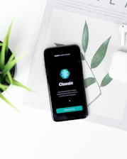 A smartphone displaying a breathing exercise app is placed on a white surface. There are green leaves in the background, partially covered by the phone. Nearby, there is a pair of white wireless earphones.