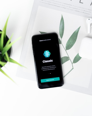 A smartphone displaying a breathing exercise app is placed on a white surface. There are green leaves in the background, partially covered by the phone. Nearby, there is a pair of white wireless earphones.