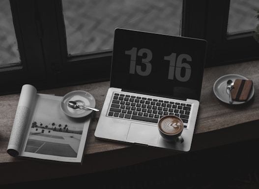 A laptop with a digital clock screensaver displays the time. Next to the laptop is a cup of cappuccino with latte art on the foam and a saucer holding a spoon. An open magazine shows a black-and-white image of a tennis court. The setting appears to be near a window with a reflective surface.