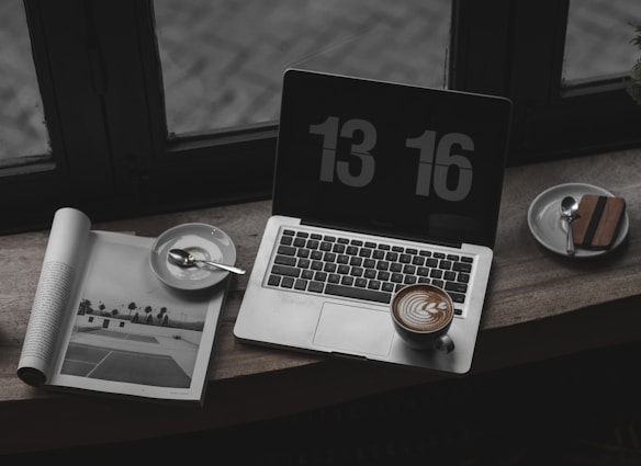 A laptop with a digital clock screensaver displays the time. Next to the laptop is a cup of cappuccino with latte art on the foam and a saucer holding a spoon. An open magazine shows a black-and-white image of a tennis court. The setting appears to be near a window with a reflective surface.