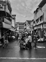 A bustling street market with numerous vendors and shoppers. The scene is filled with people engaged in various activities, including selling and purchasing goods. Surrounding buildings look weathered and closely packed, providing an enclosed feeling to the market scene. The sky is overcast, contributing to the overall atmosphere.