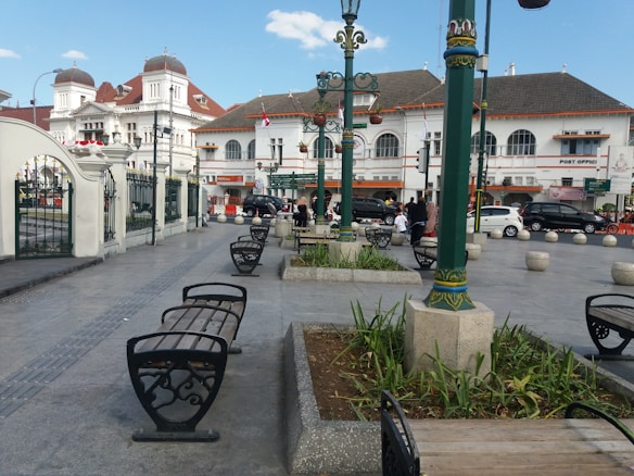 A public square with benches, street lamps, and a historic-looking white building with a post office. The area is paved with concrete and adorned with small green plants in planters. Several people are walking and interacting in the background, and cars are parked along the street.