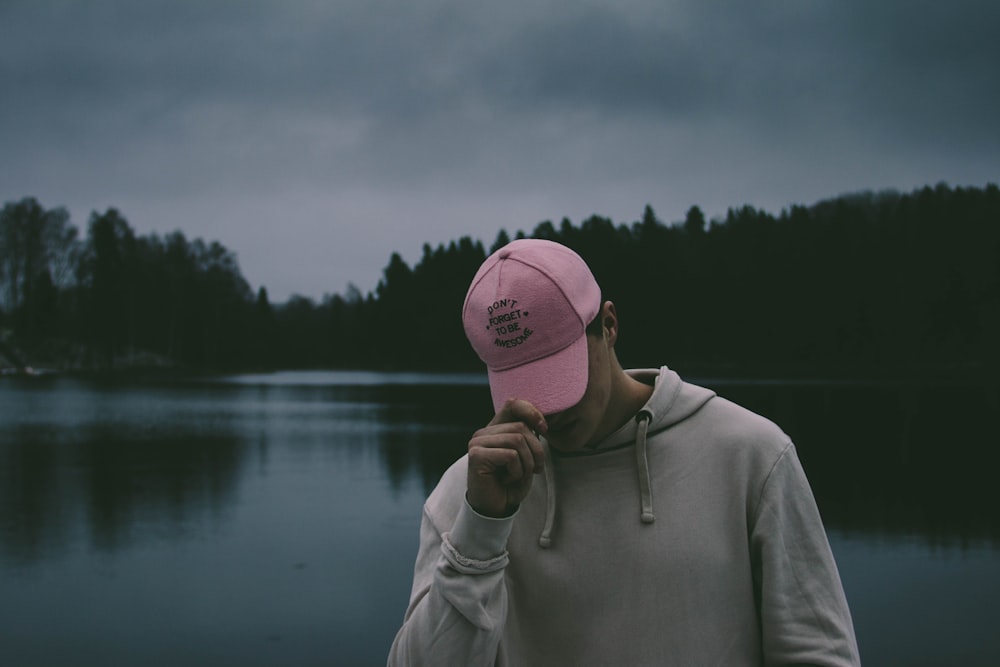 man wearing white pullover hoodie holding his pink cap overlooking body of water